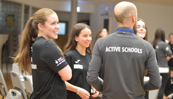 Active Schools staff and students chat in a bright school hallway, smiling and sharing a friendly, welcoming moment at an event. 