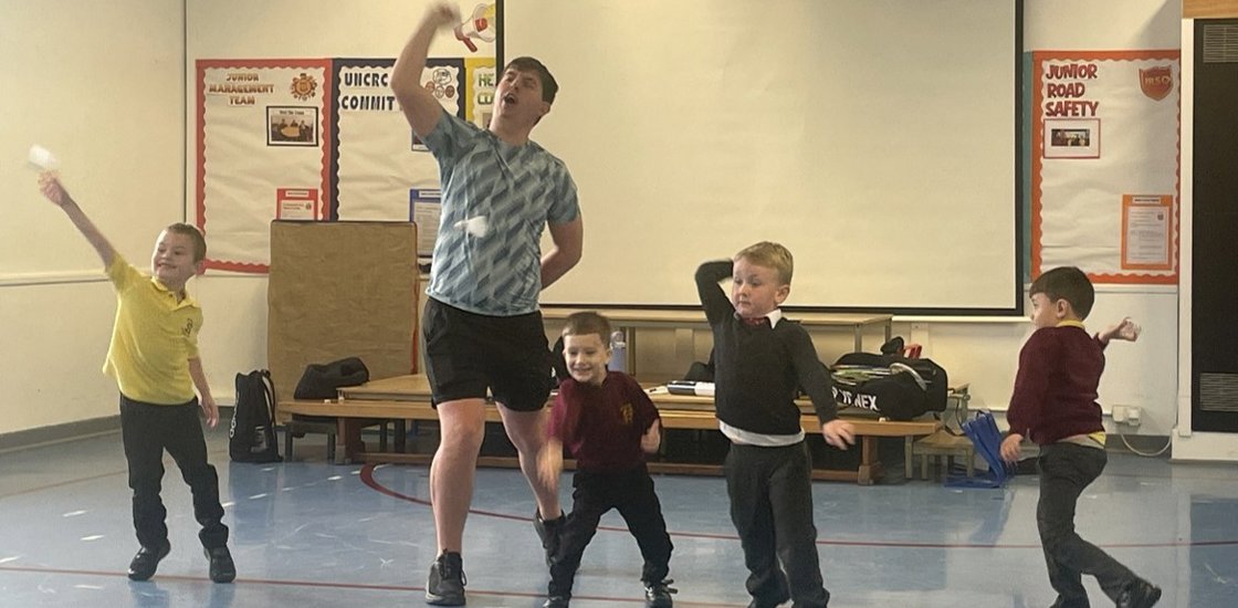 A coach and four children practice playful movement poses in a school gym, raising arms and balancing; the class feels energetic and cheerful.