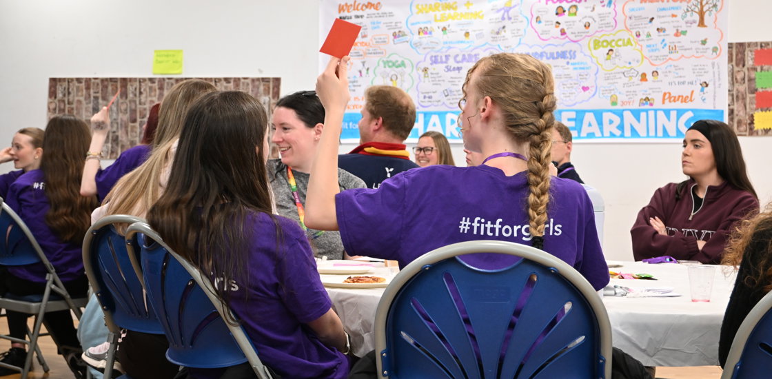 Students in purple "Fit for Girls" shirts gather around classroom tables, raising hands during a lively, collaborative workshop beneath colorful posters and brainstorming displays.