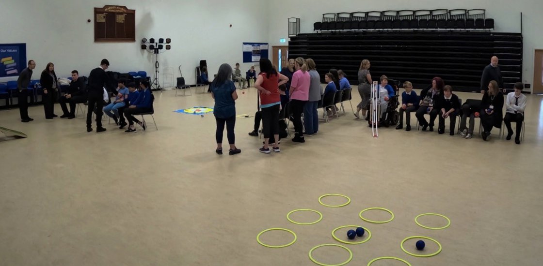 In a school gym, adults and children gather near seated spectators while small balls sit among bright floor hoops, creating a relaxed activity scene.