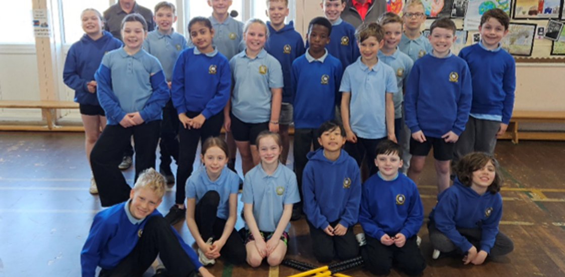 Schoolchildren and two adults pose smiling in blue uniforms in a classroom gym, with red-and-yellow sports equipment.
